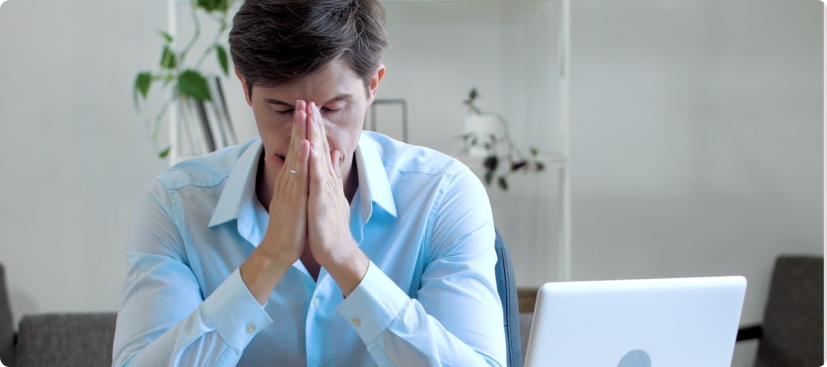 A man at a desk with a laptop at his side, and his hands pressed together on his nose in frustration.